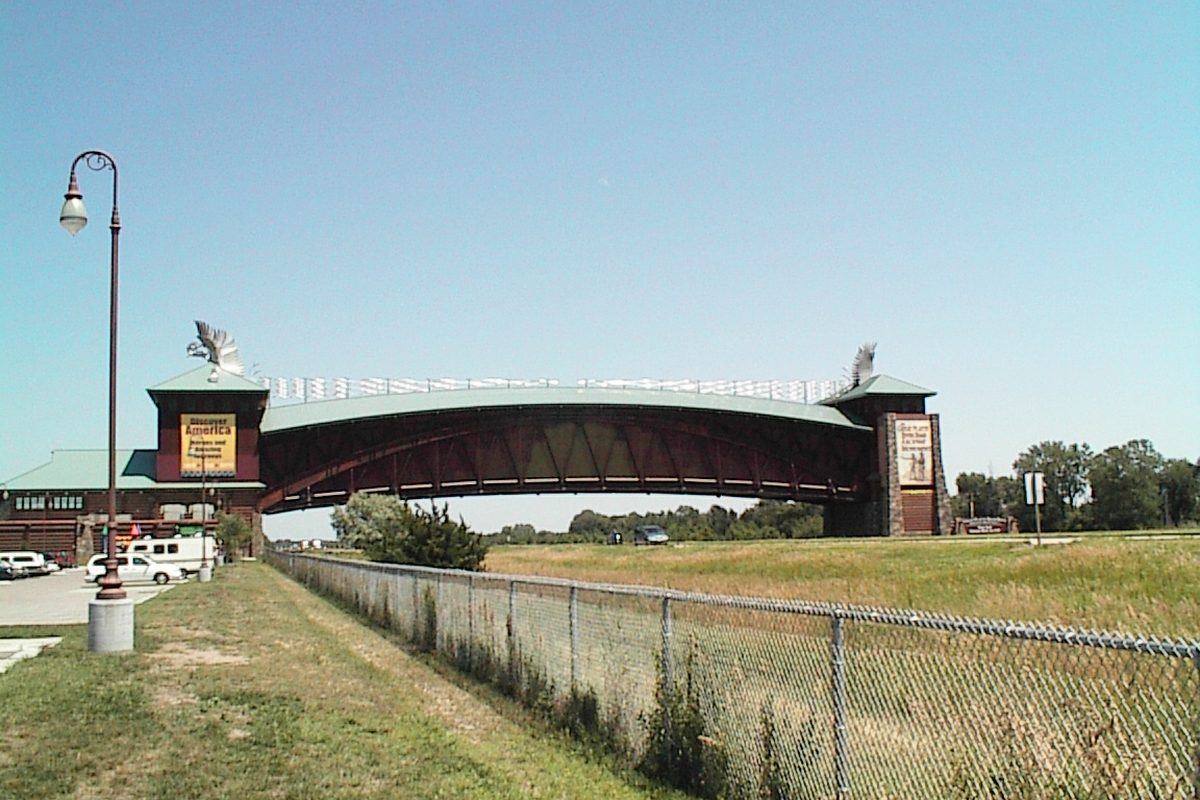 The Great Platte River Road Archway near Kearney, a dark red arch with green roofs spanning the highway, seen past a chain-link fence and a grassy field