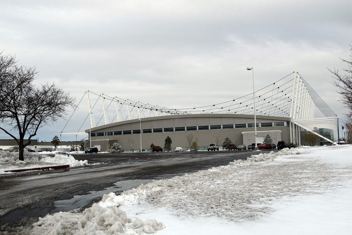 The Utah Olympic Oval in Kearns, a long arena hung from white cable masts, with plowed snowbanks and a parking lot in front