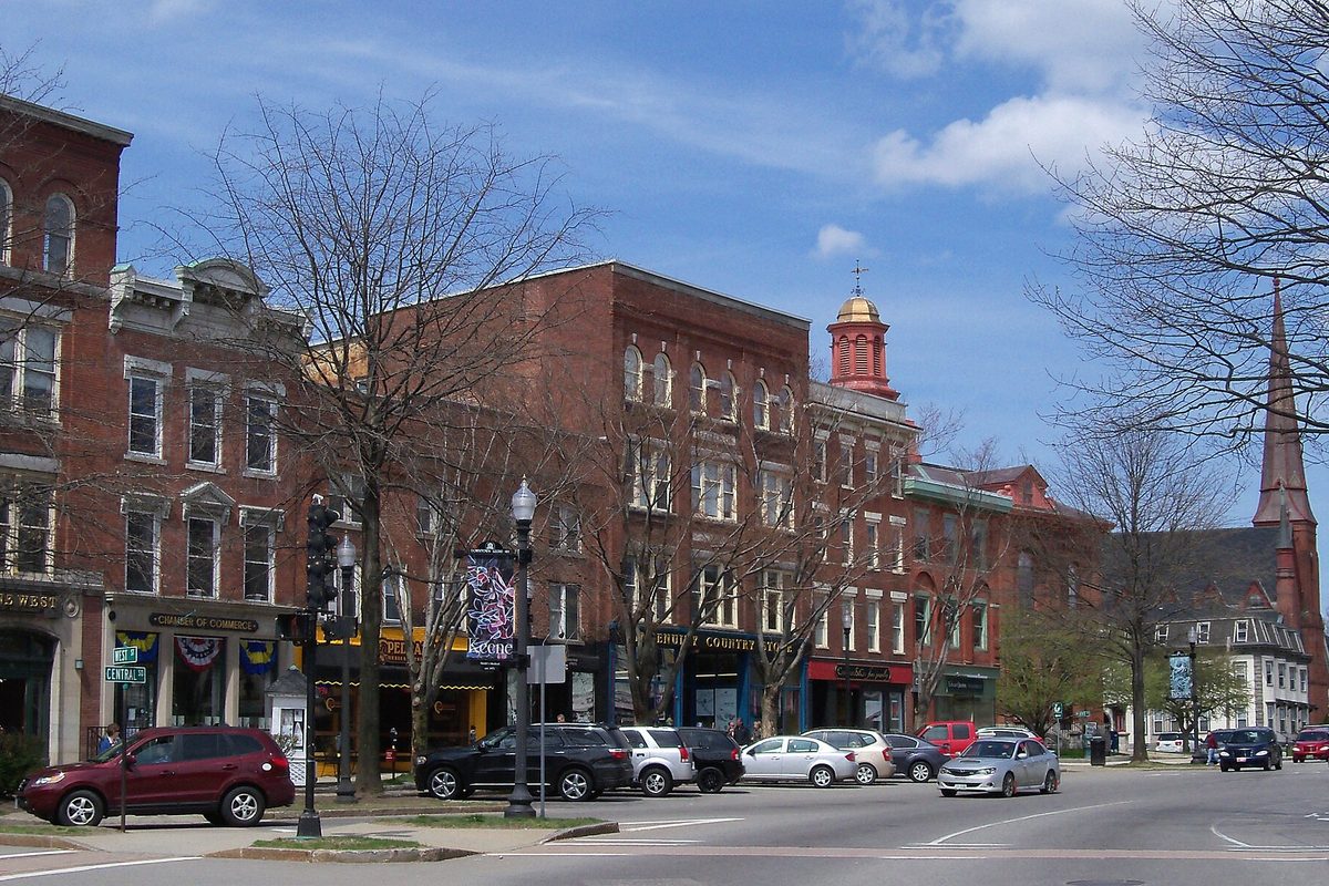 Main Street in downtown Keene, a row of red-brick commercial buildings with a red cupola and a tall church steeple, cars parked along the street and bare trees in early spring