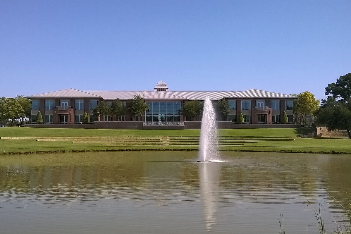 Keller City Hall, a long two-story building with a small dome, seen across a pond with a tall fountain and a terraced lawn