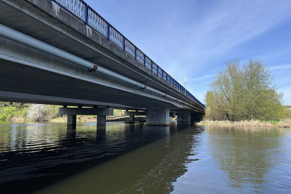A concrete road bridge with blue railings crossing the calm Sammamish River in Kenmore, seen from water level on a sunny spring day
