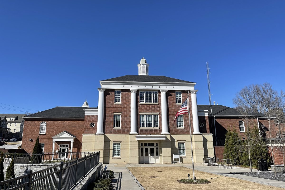 Red-brick Kennesaw City Hall with tall white columns, a white cupola and an American flag, under a clear blue sky