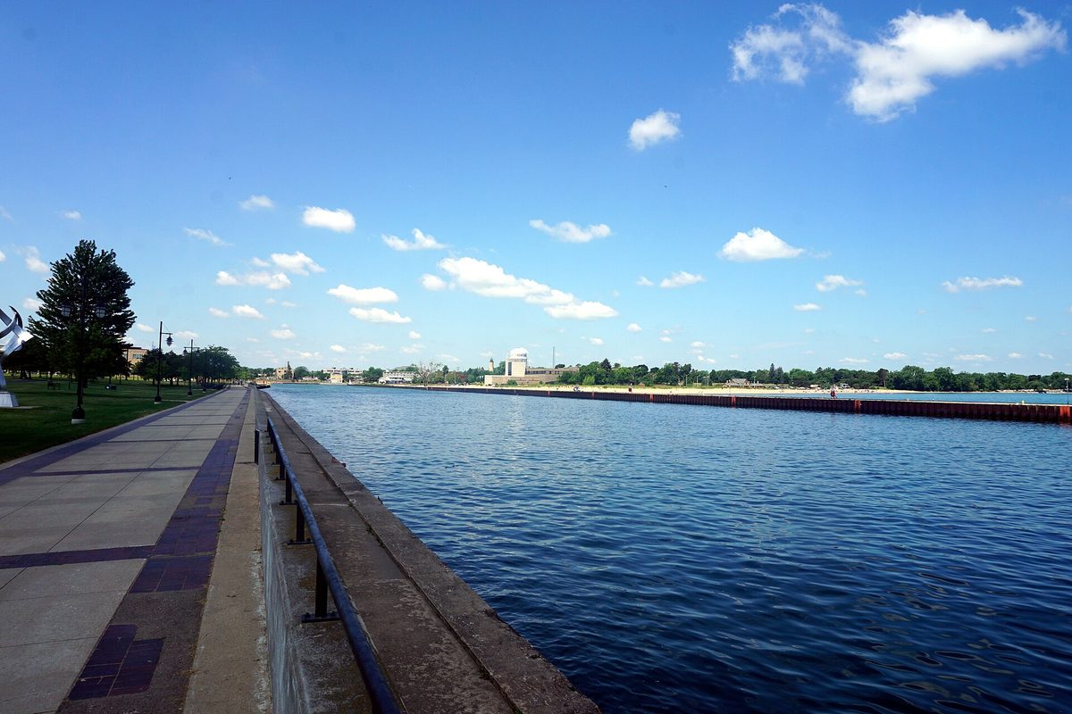 A paved walkway and railing beside the Pike River Canal in Kenosha, with calm blue water and a long breakwater under a blue sky