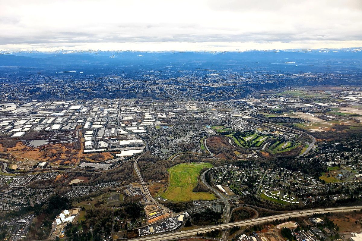 Aerial view of the Kent valley under a cloudy sky, with rows of warehouse roofs, a golf course, a freeway and mountains in the distance