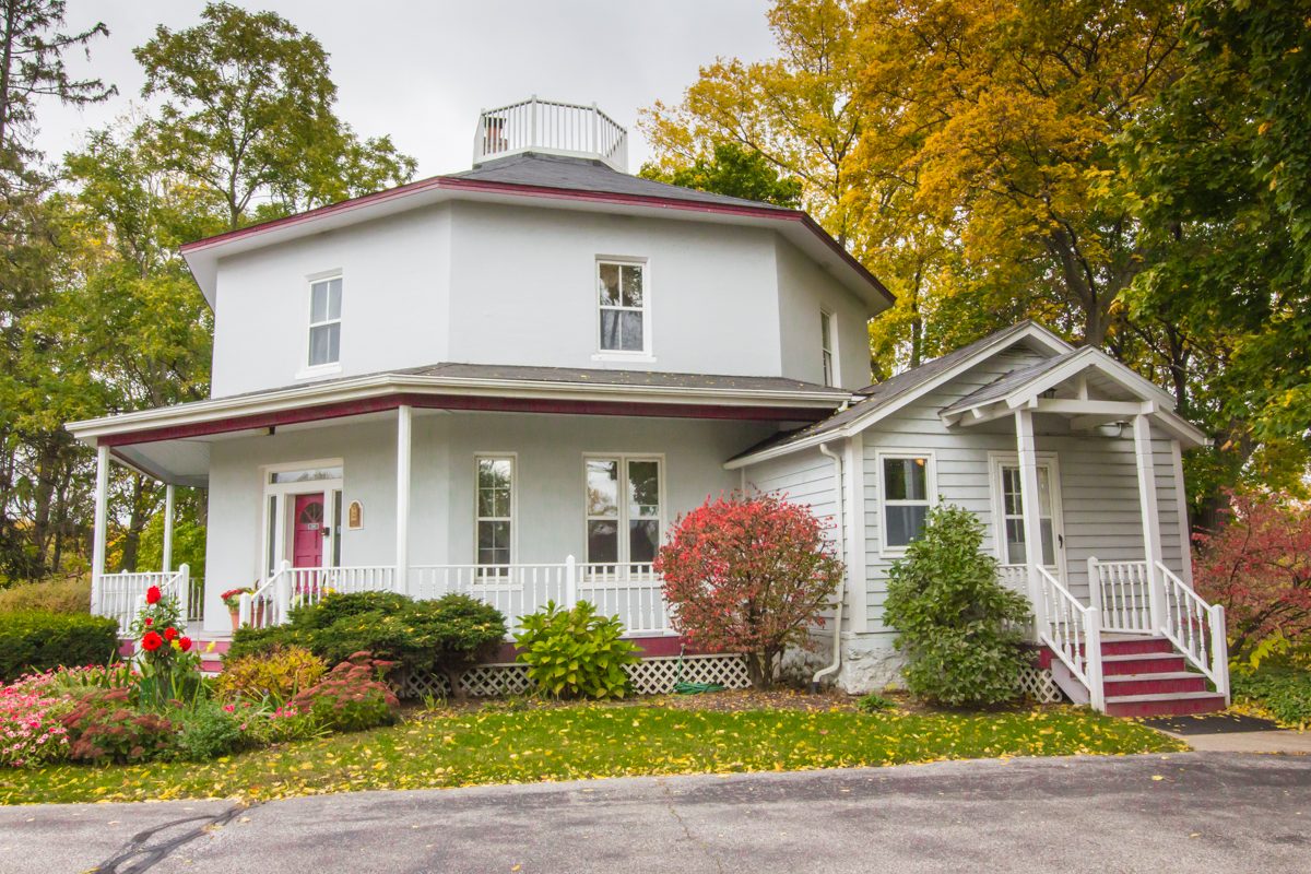 The white octagonal Bailey House in Kentwood with a red front door, a railed porch and autumn trees behind it