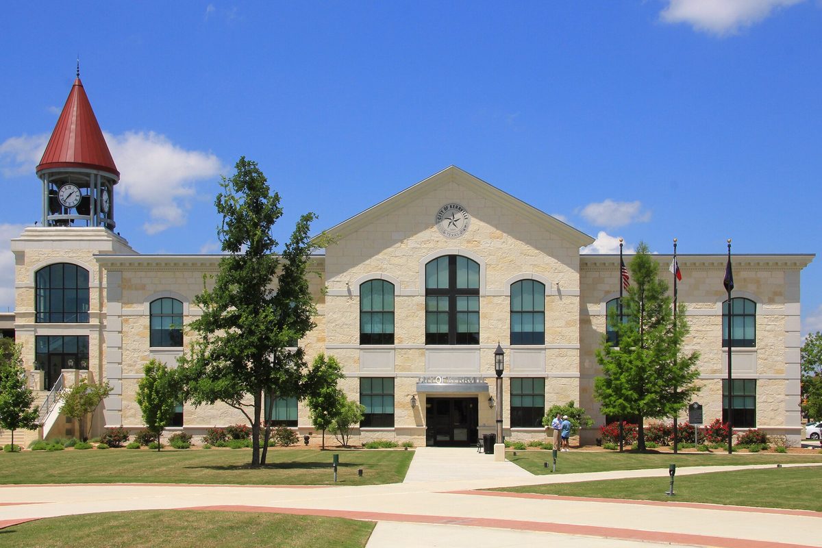 Kerrville City Hall, a pale limestone building with tall arched windows and a red-roofed clock tower, behind a lawn, young trees and flags