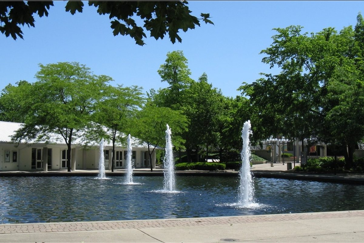 Four fountain jets rising from a pool in front of the low white administration building at Fraze Pavilion in Kettering, framed by leafy trees