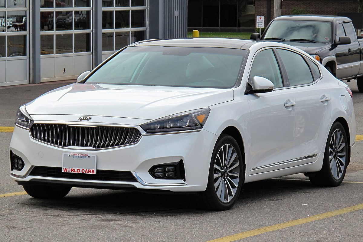 A white 2017 Kia Cadenza sedan on a dealer lot, seen from the front left, with a pickup truck parked behind it