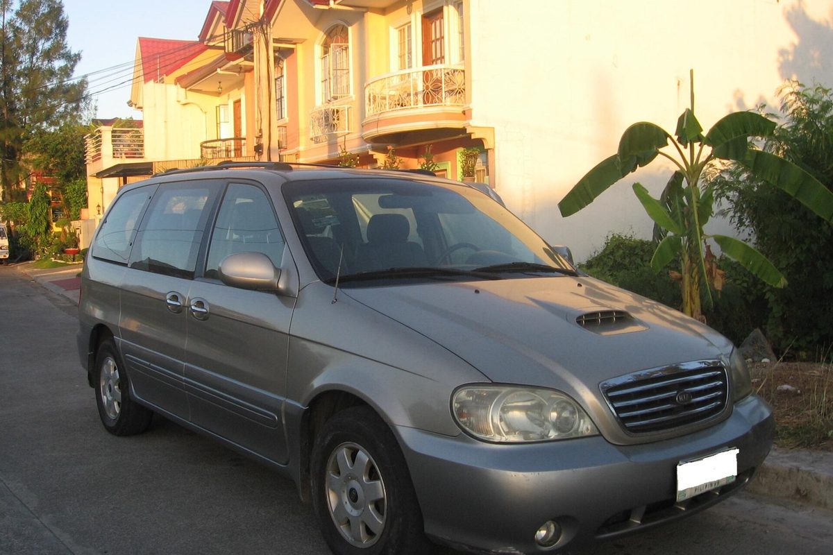 A silver early-2000s Kia Carnival minivan parked on a residential street beside a banana plant