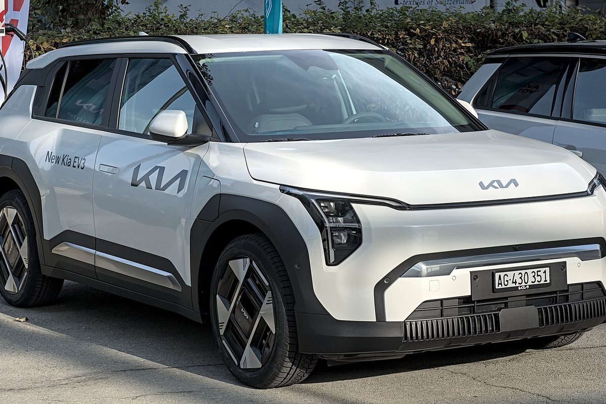 A white Kia EV3 compact electric SUV with black wheel arches and a New Kia EV3 door decal, at an auto show, seen from the front right