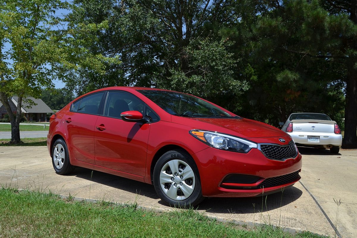 A red 2015 Kia Forte sedan parked on a concrete driveway next to a lawn, with tall pine trees behind it