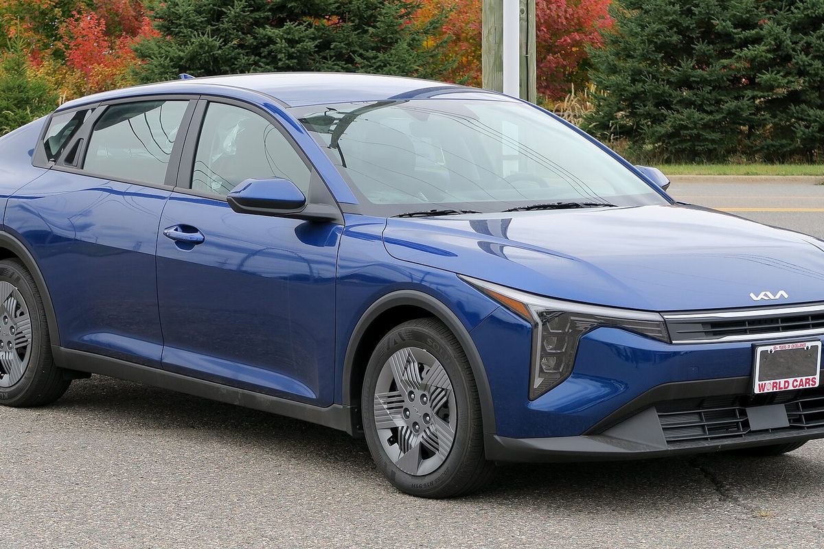 A blue 2025 Kia K4 sedan parked on asphalt, seen from the front right, with autumn trees behind it