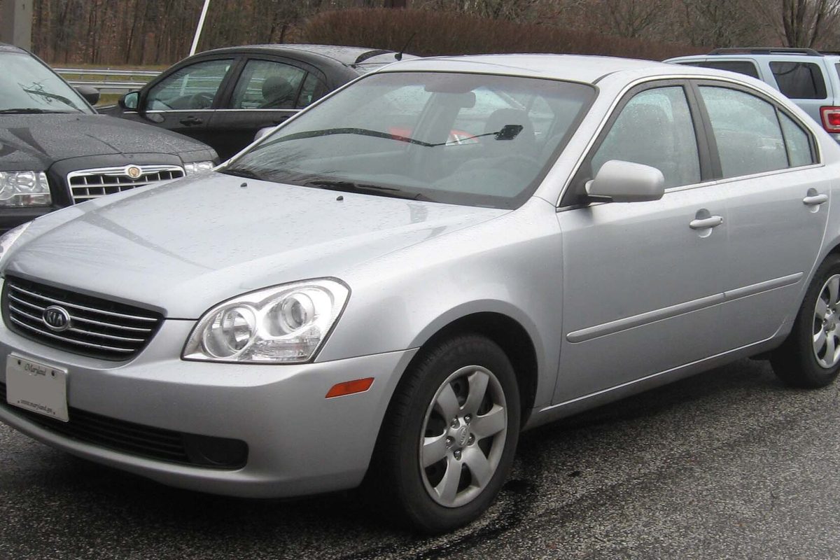 A silver 2006-2008 Kia Optima sedan with five-spoke alloy wheels, parked on wet pavement in a lot