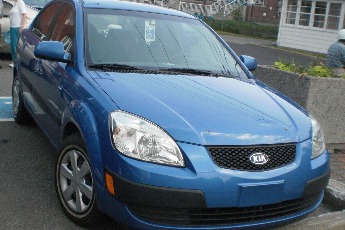 A bright blue second-generation Kia Rio sedan parked at a curb beside a concrete planter