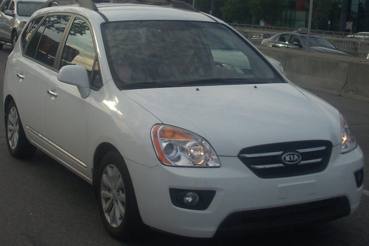 A white Kia Rondo wagon driving on a city road, front three-quarter view