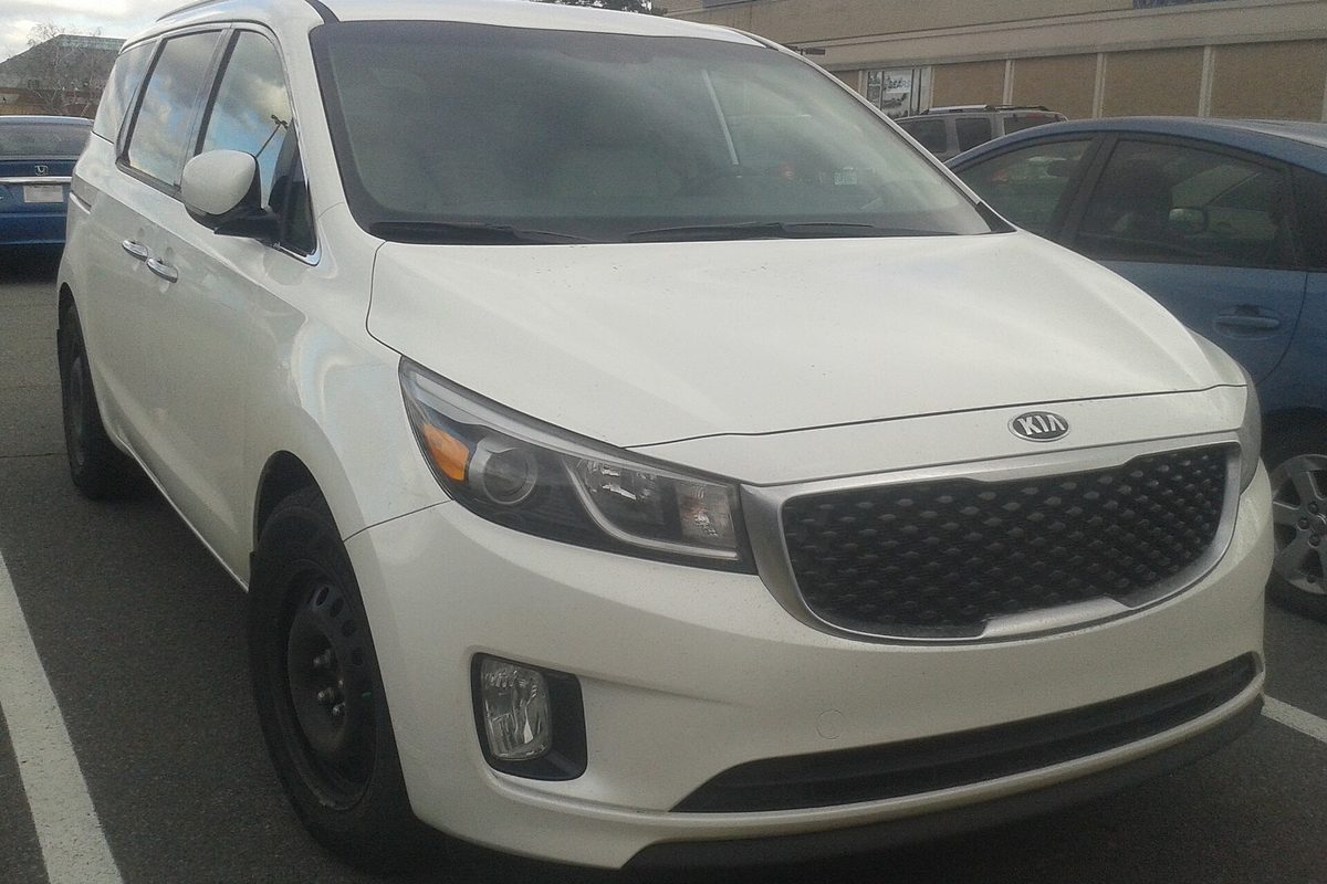 A white 2015 Kia Sedona minivan on black steel wheels, parked in a lot on an overcast day
