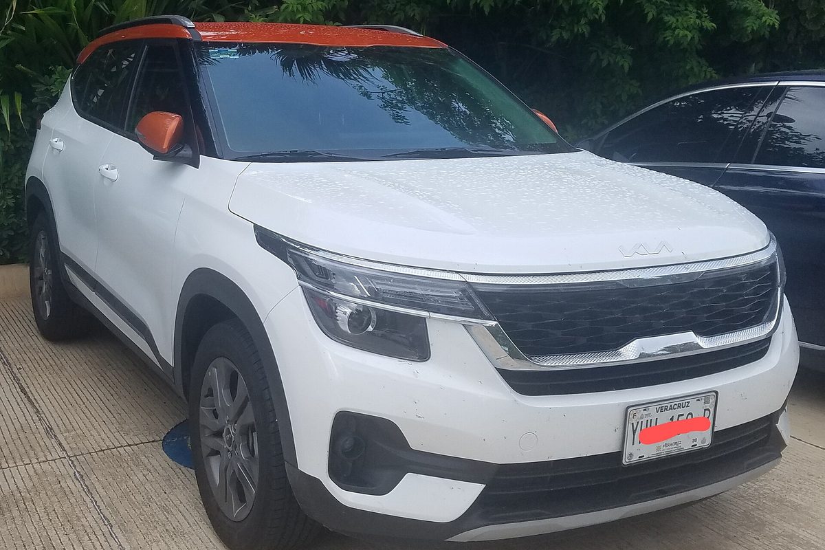 A white Kia Seltos with an orange roof and mirror caps, parked on wet pavement in front of trees, plate blurred