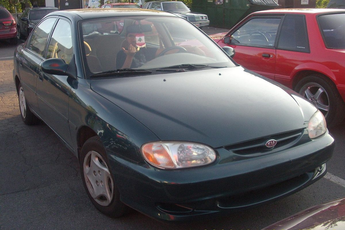 A dark green Kia Sephia sedan parked in a lot beside a red coupe, a passenger visible through the windshield