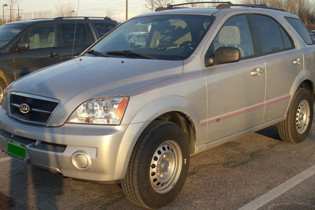 A silver 2003-2006 Kia Sorento SUV on steel wheels with a thin red side stripe, parked in a lot beside a dark SUV
