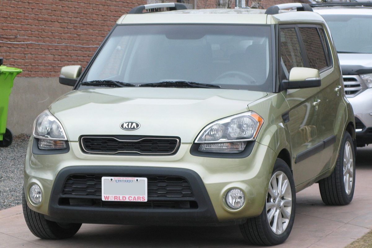 A pale green 2013 Kia Soul with a roof rack and fog lights, parked in front of a brick wall