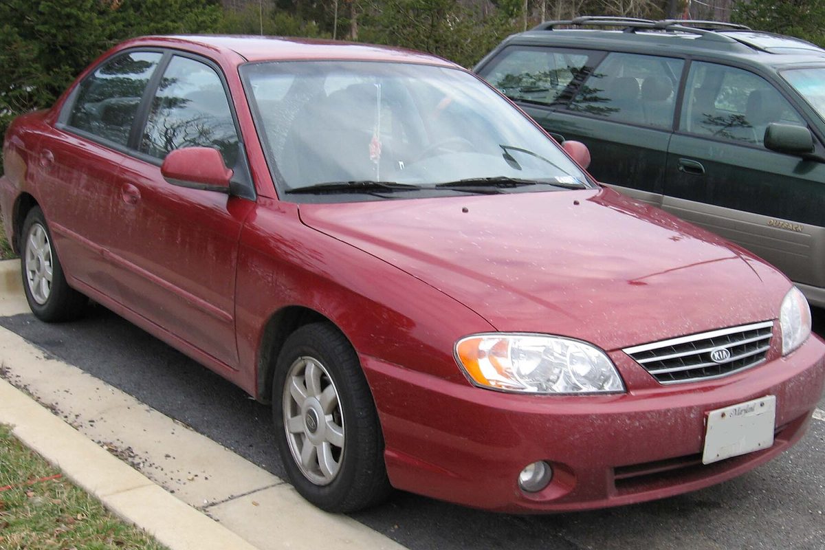 A red first-generation Kia Spectra sedan with road grime on the paint, parked at a curb with an SUV behind it