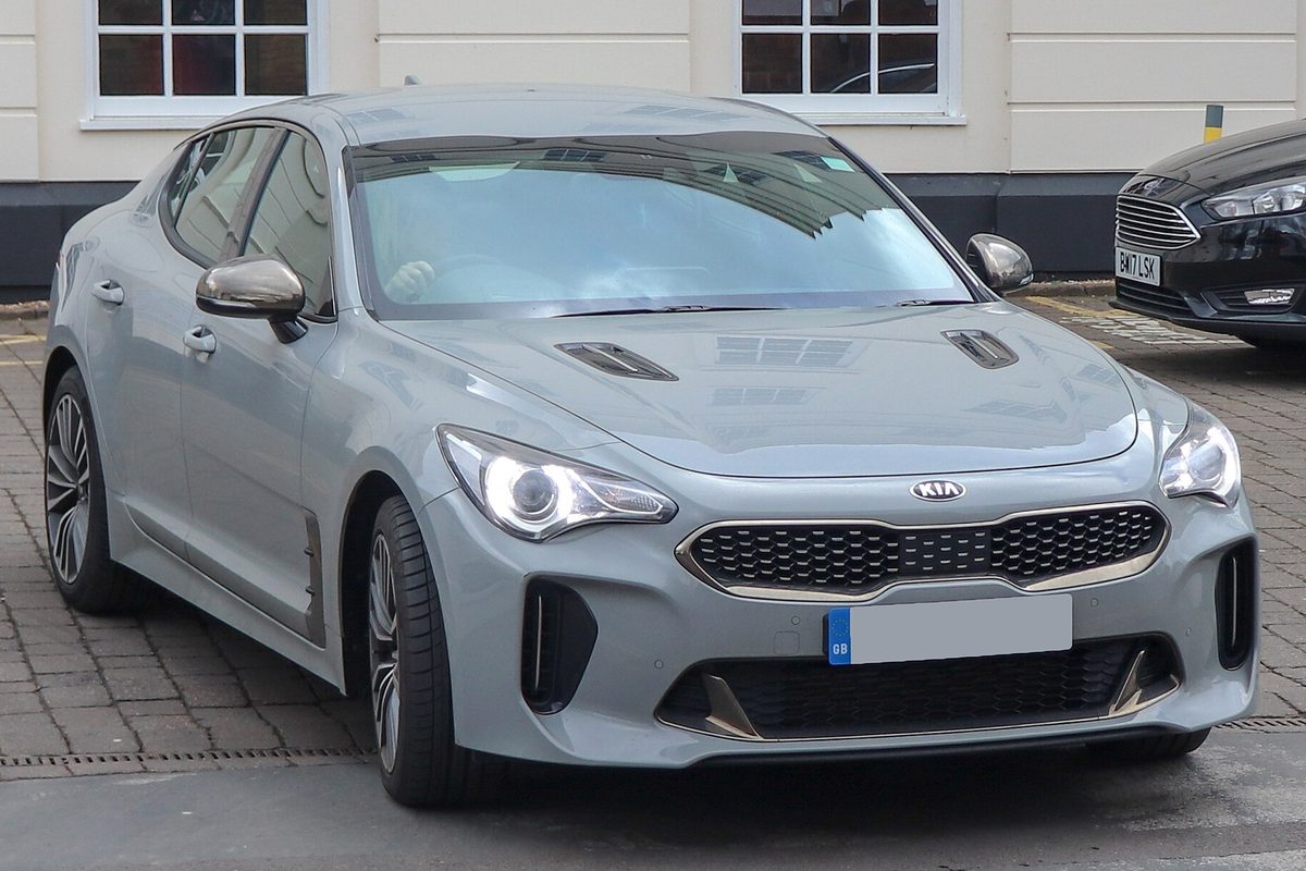 A light gray Kia Stinger fastback parked on a brick-paved street, seen from the front right, with a white building behind it