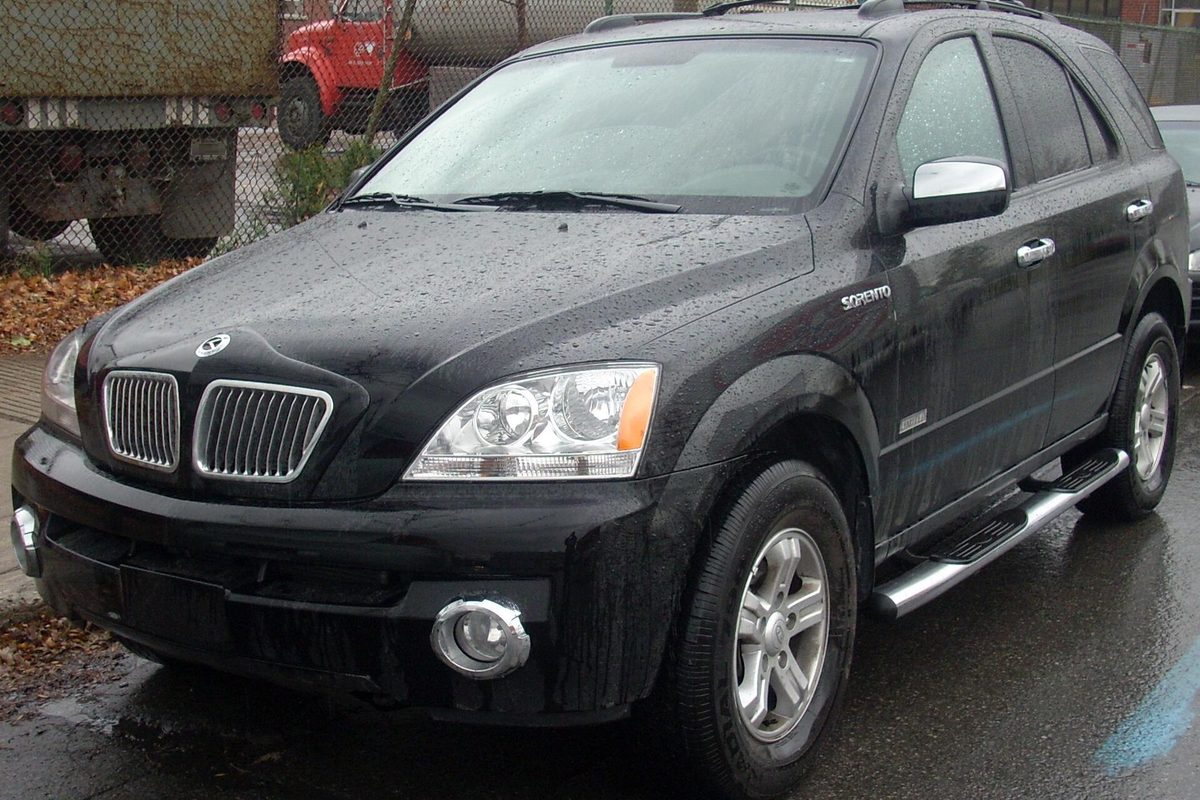 A black first-generation Kia Sorento SUV, 2003-2006 style, parked on a rain-wet street beside a chain-link fence, photographed in Montreal