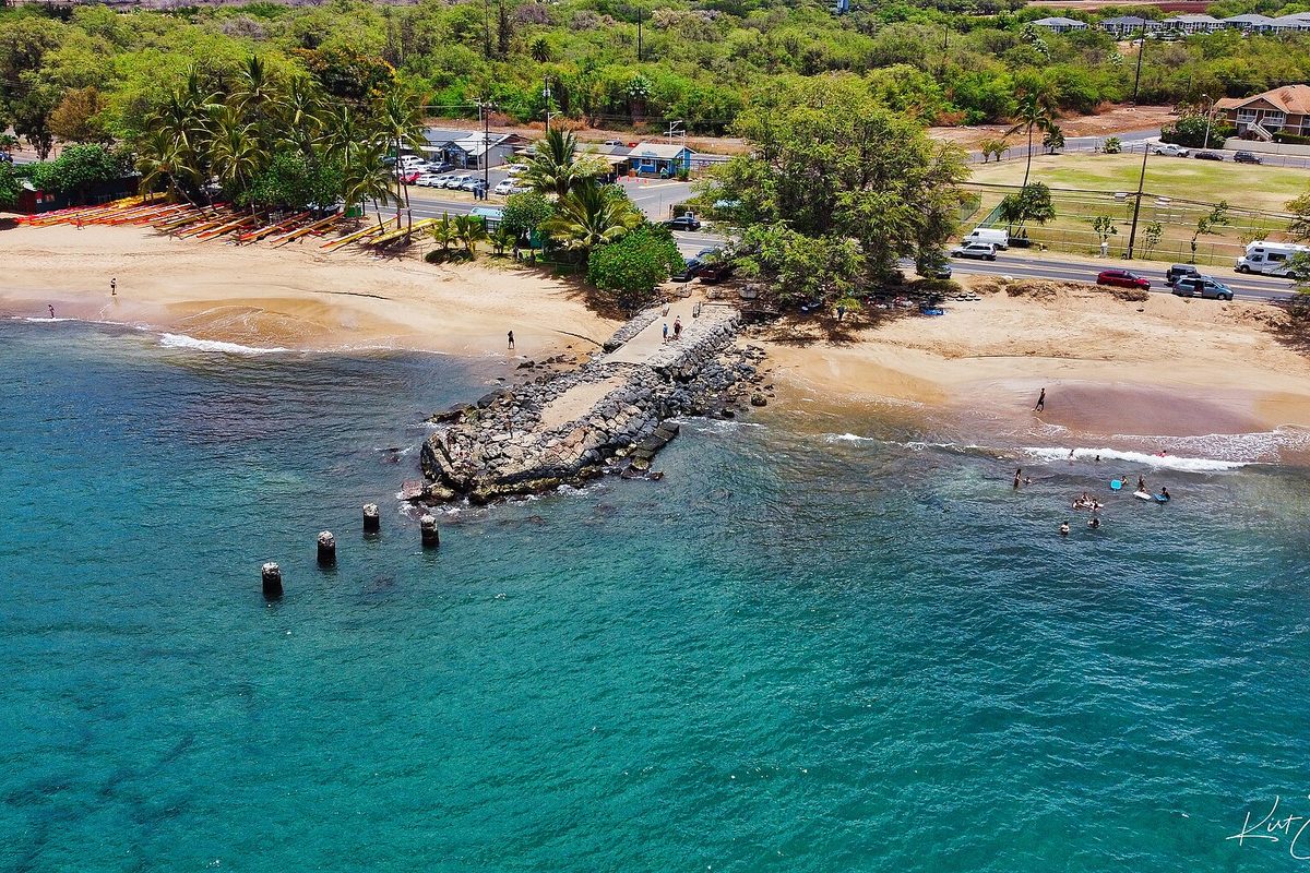 Aerial view of the old stone Kihei Wharf reaching into turquoise water, with a sandy beach, swimmers, palm trees and cars parked along the coast road