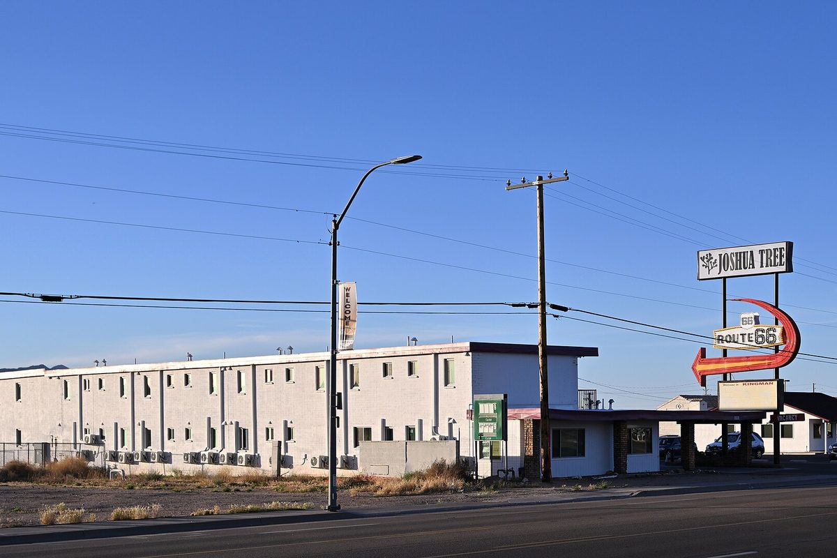 A white two-story former motel, now the Joshua Tree apartments, beside a vintage Route 66 arrow sign under a clear blue Kingman sky