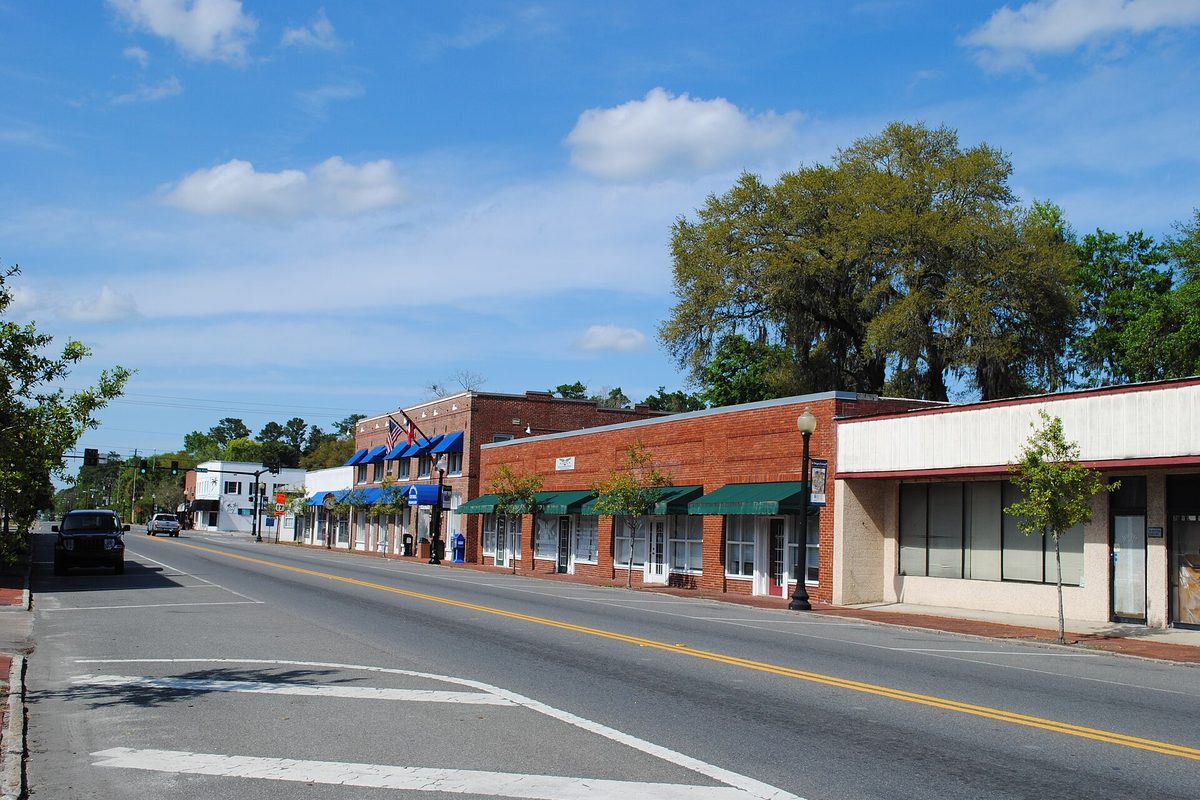 A quiet downtown Kingsland street lined with red-brick storefronts under green awnings, a large oak tree behind them and a clear blue sky