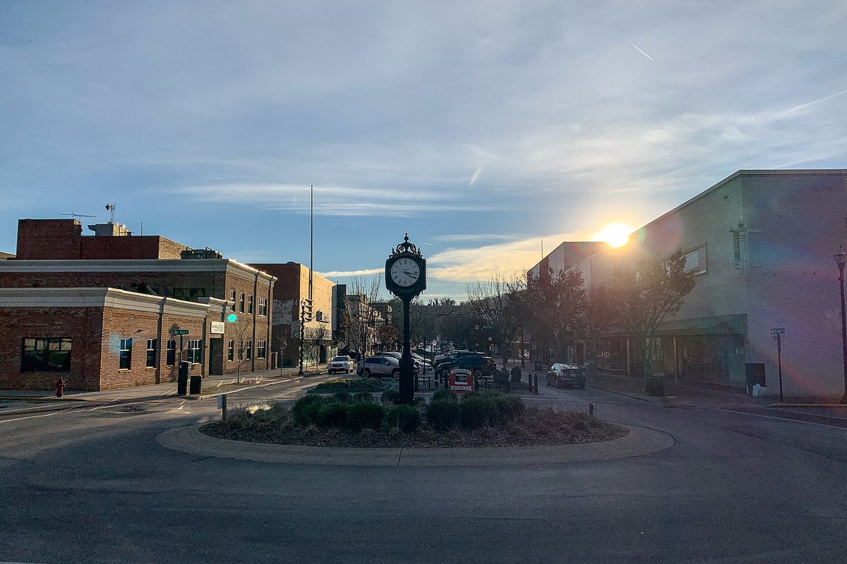 A street clock in a planted roundabout on Broad Street in downtown Kingsport, with the low sun behind brick storefronts