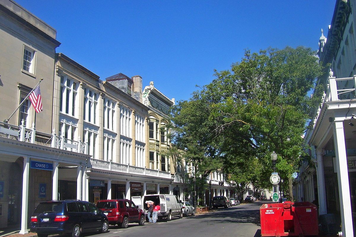 A street in Kingston's Stockade District lined with pale historic buildings with balconies and columned porches, parked cars and a large shade tree