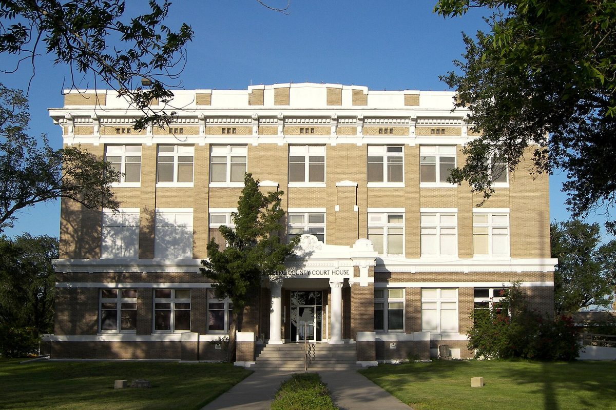 The four-story tan brick Kleberg County Courthouse in Kingsville with a white columned entrance, framed by trees and a green lawn under a blue sky