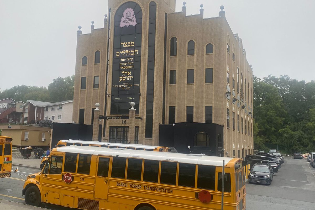 A tall tan stone building with a large Hebrew-lettered panel on its front in Kiryas Joel, with yellow school buses parked on the street before it