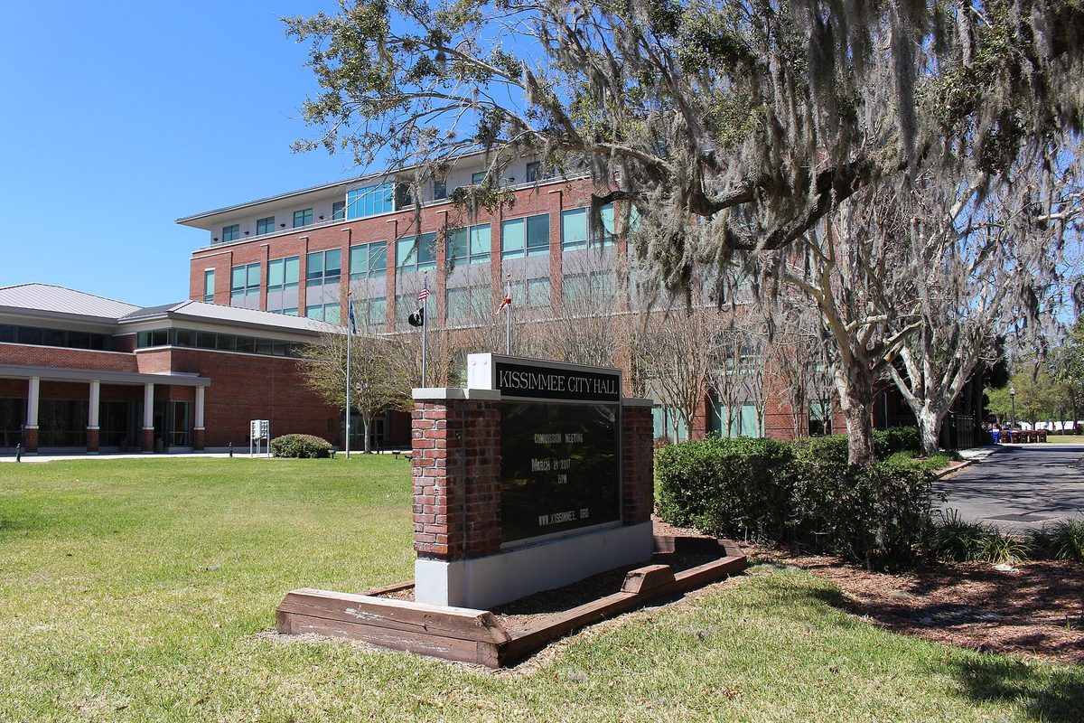 A brick Kissimmee City Hall sign on the lawn in front of the brick and glass city hall building, with a moss-draped oak at right