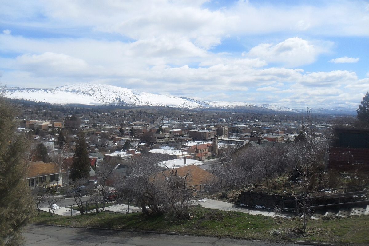 Downtown Klamath Falls seen from a hillside street, rooftops and bare trees below snow-covered mountains under a cloudy sky