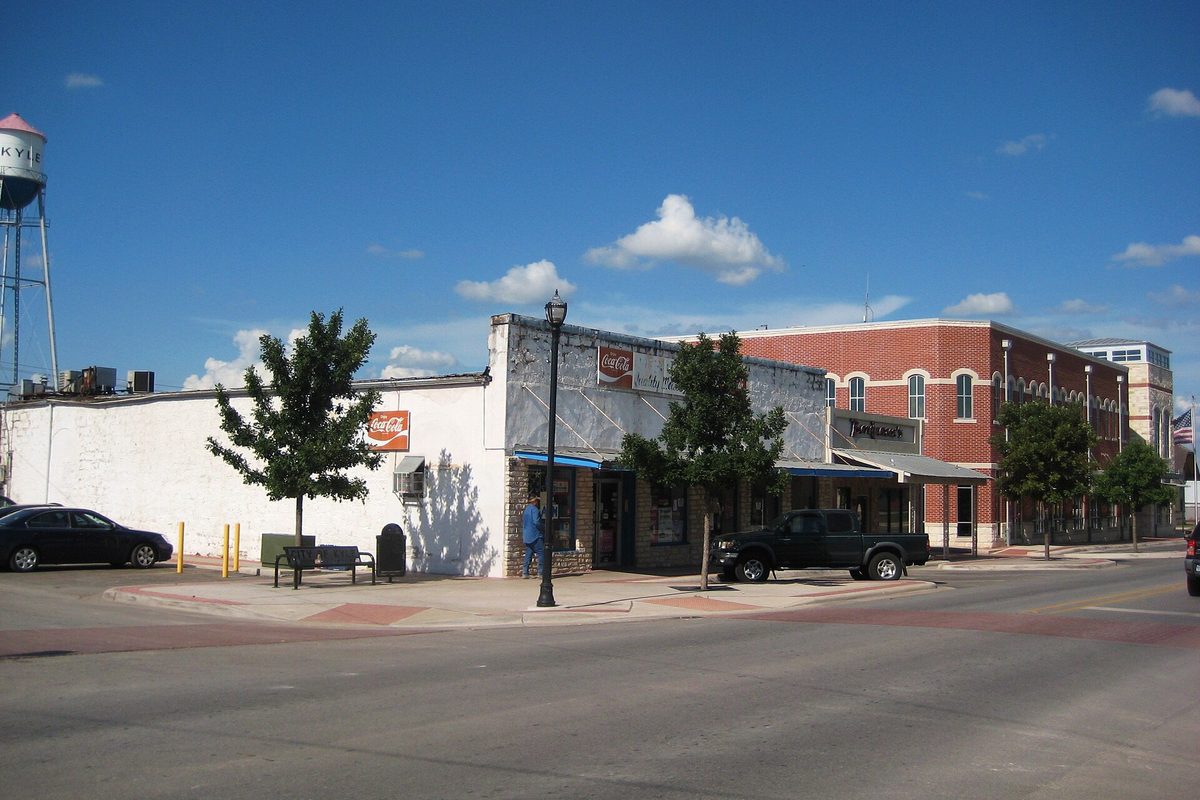 A street corner in downtown Kyle with a white storefront, a red brick building and a pickup truck parked at the curb, and the KYLE water tower against a blue sky
