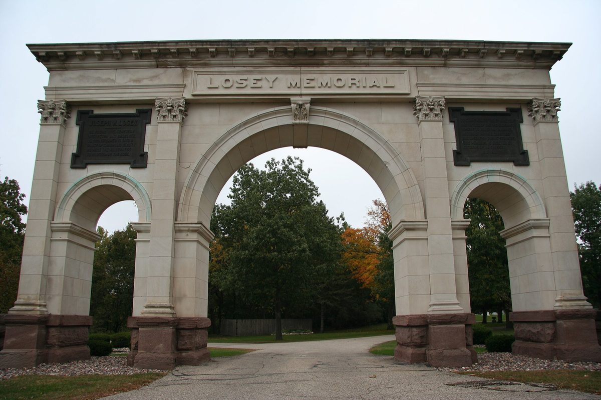 The pale stone Losey Memorial Arch, three arched openings over a paved drive at the entrance to Oak Grove Cemetery, with autumn trees behind it