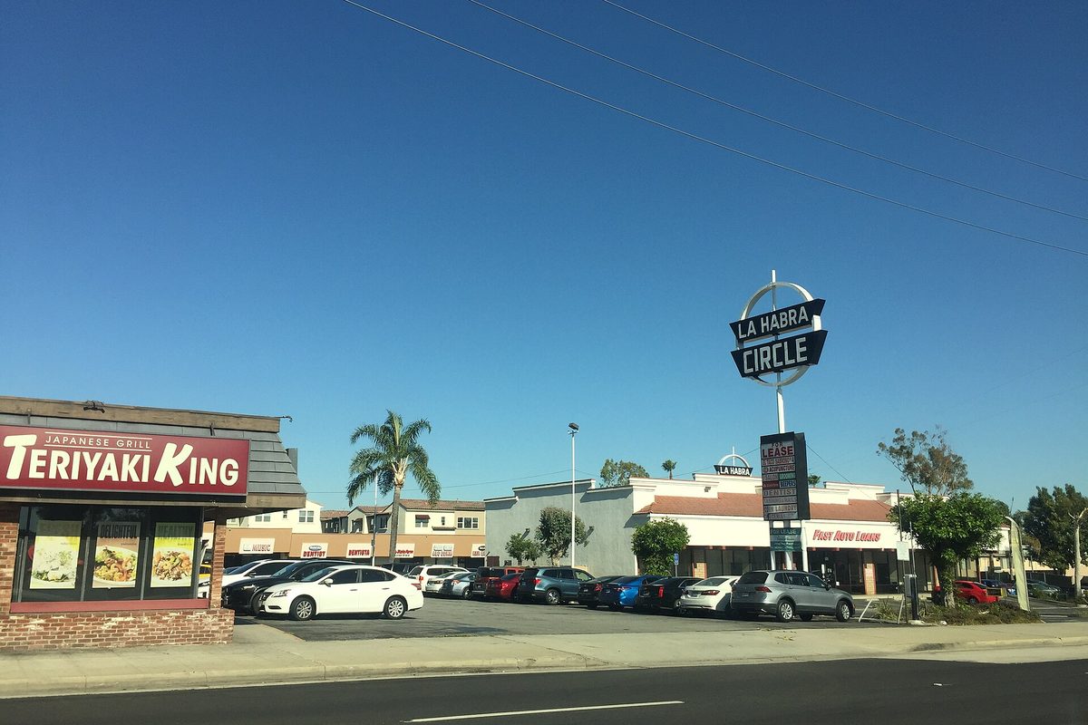 The mid-century La Habra Circle sign towering over a parking lot at the shopping center, with palm trees and a clear blue sky