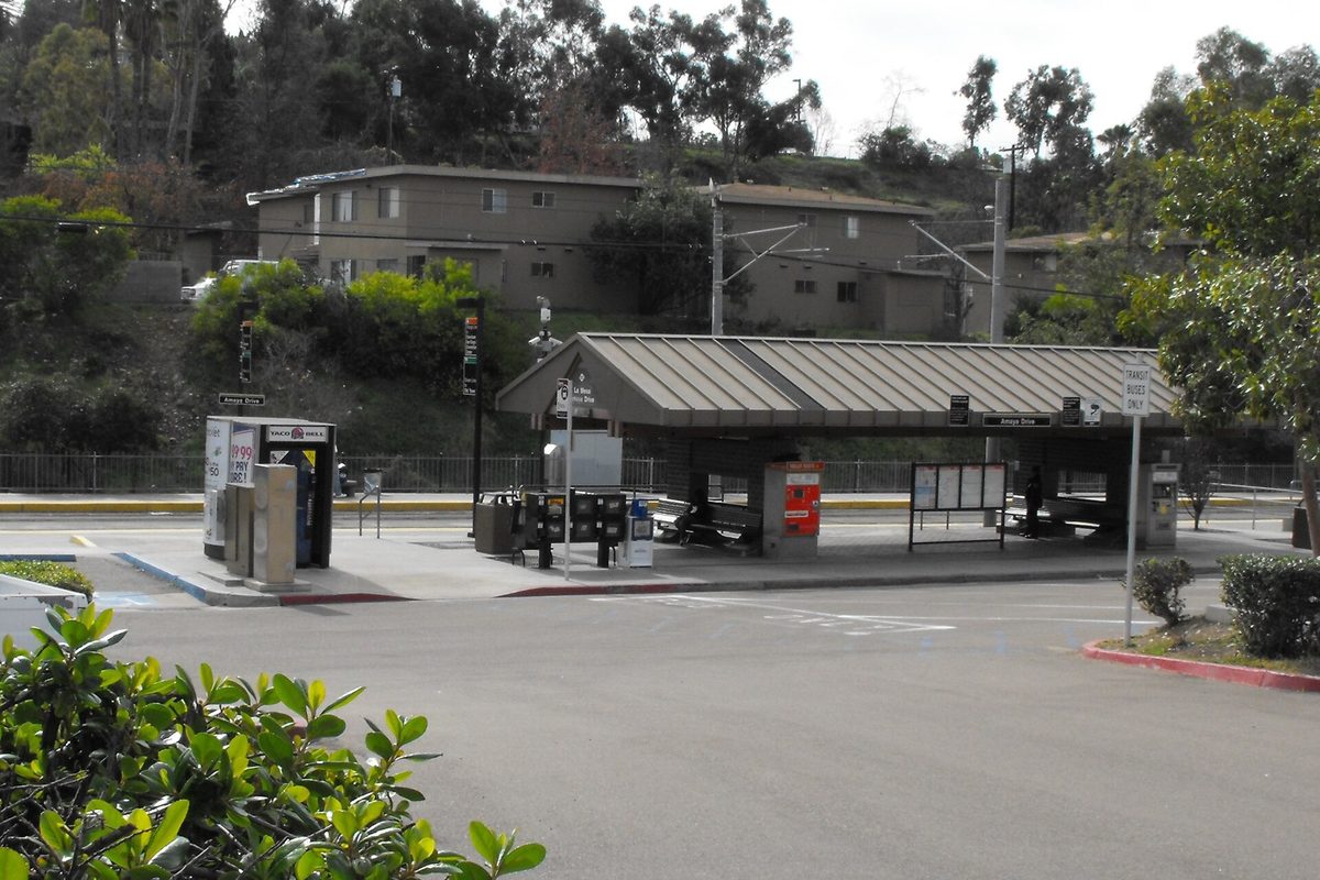 The covered platform at the Amaya Drive trolley station in La Mesa, with ticket machines, an empty parking lot and apartments on the hillside behind