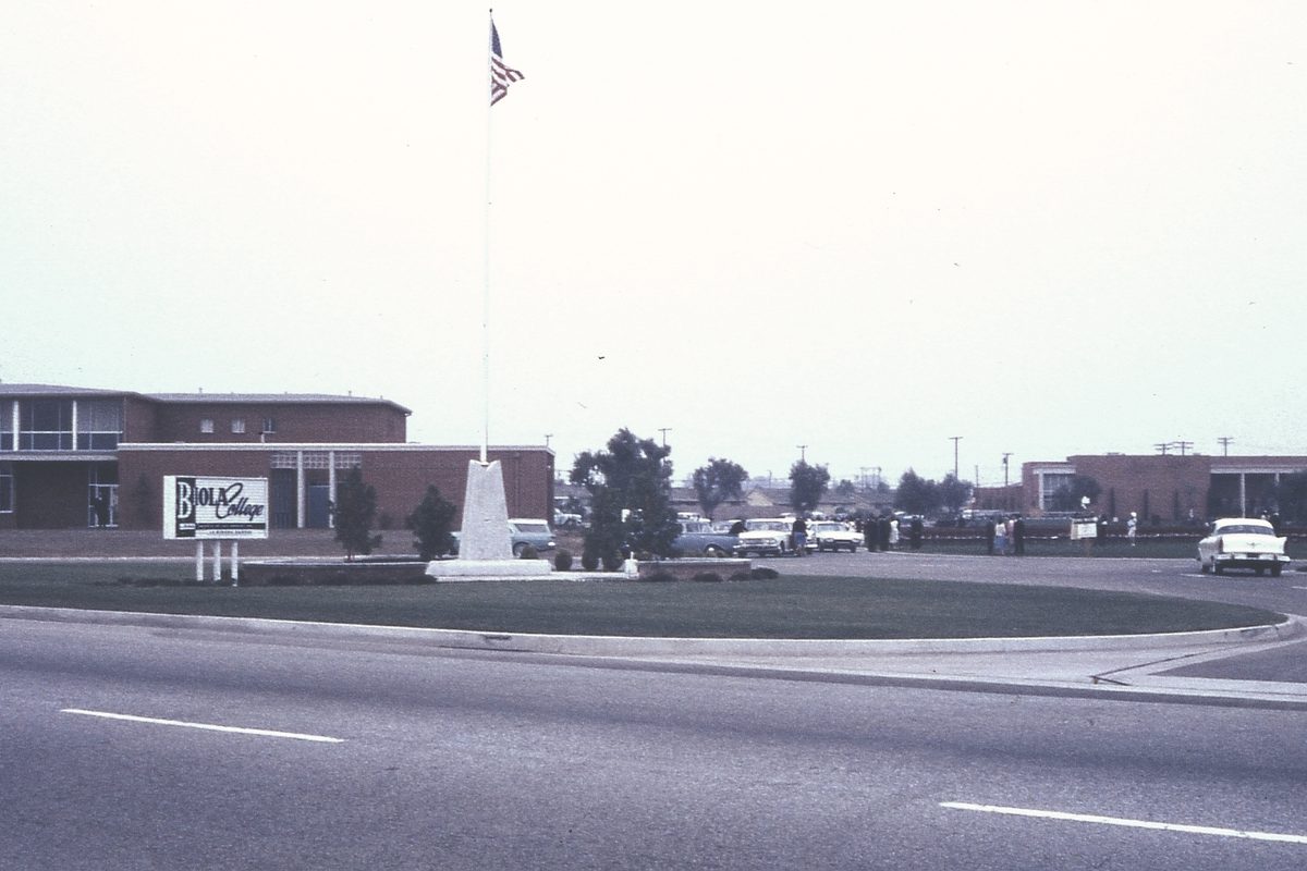 A 1963 color slide of the Biola College entrance in La Mirada, with the college sign, a flagpole and a low brick building beyond a landscaped median, and 1960s cars parked nearby