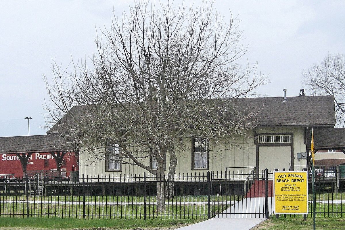 The Old Sylvan Beach Depot in La Porte behind a black iron fence, with a bare tree in front, a yellow heritage society sign and a red Southern Pacific car to the left