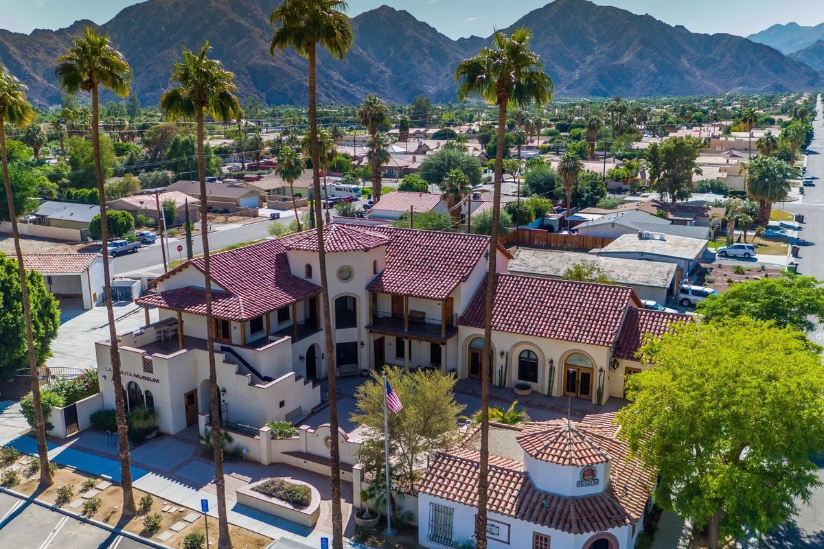 Aerial view of the La Quinta Museum, a white Spanish-style building with red tile roofs and palms, with rocky desert mountains behind