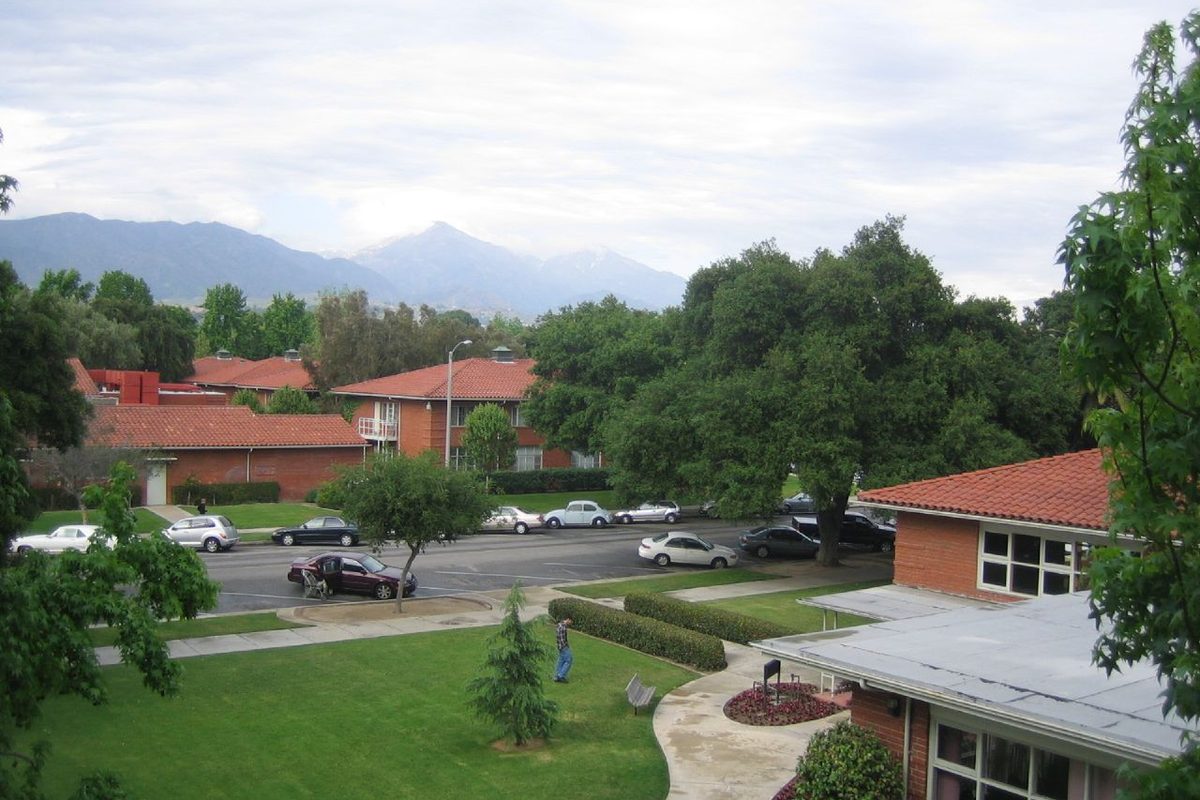 Red brick buildings with tile roofs along Third Street at the University of La Verne, with lawns, oak trees, a row of parked cars and mountains under gray clouds