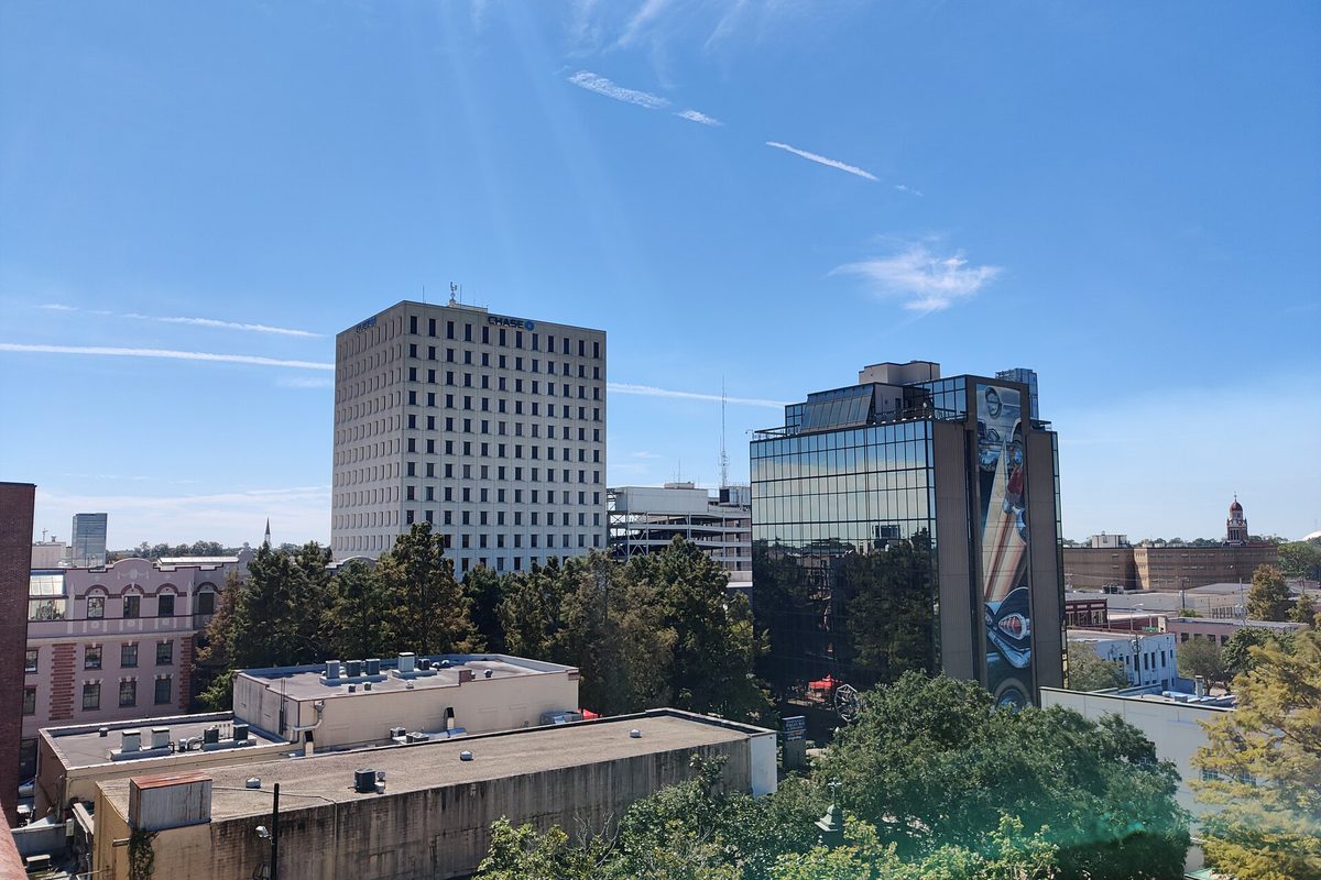 Downtown Lafayette office towers, one with a tall mural on its side, above trees and low rooftops on a sunny day