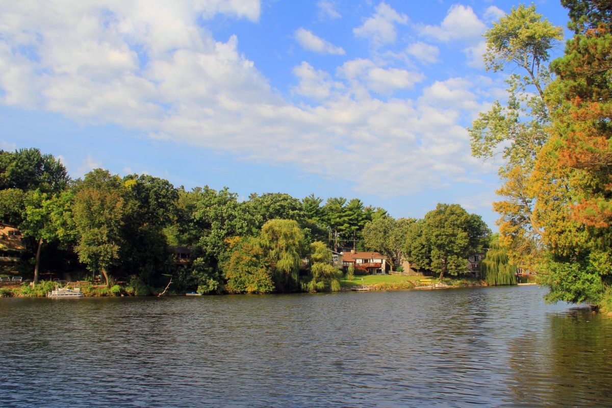 Woods Creek Lake in Lake in the Hills, with a wooded shore, a red-roofed house among the trees and a small boat at a dock under a partly cloudy sky