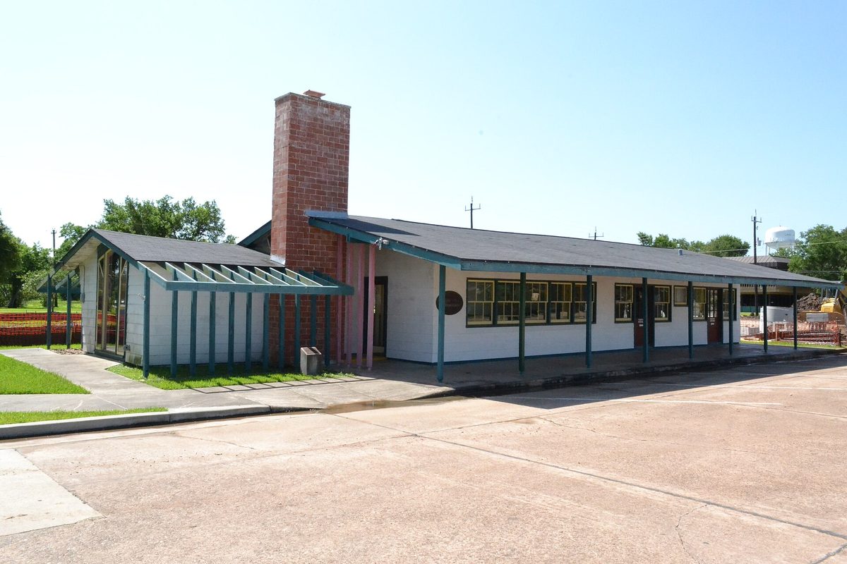 The low white Alden B. Dow office building in Lake Jackson, with teal porch posts, a tall brick chimney and a sunny parking lot in front