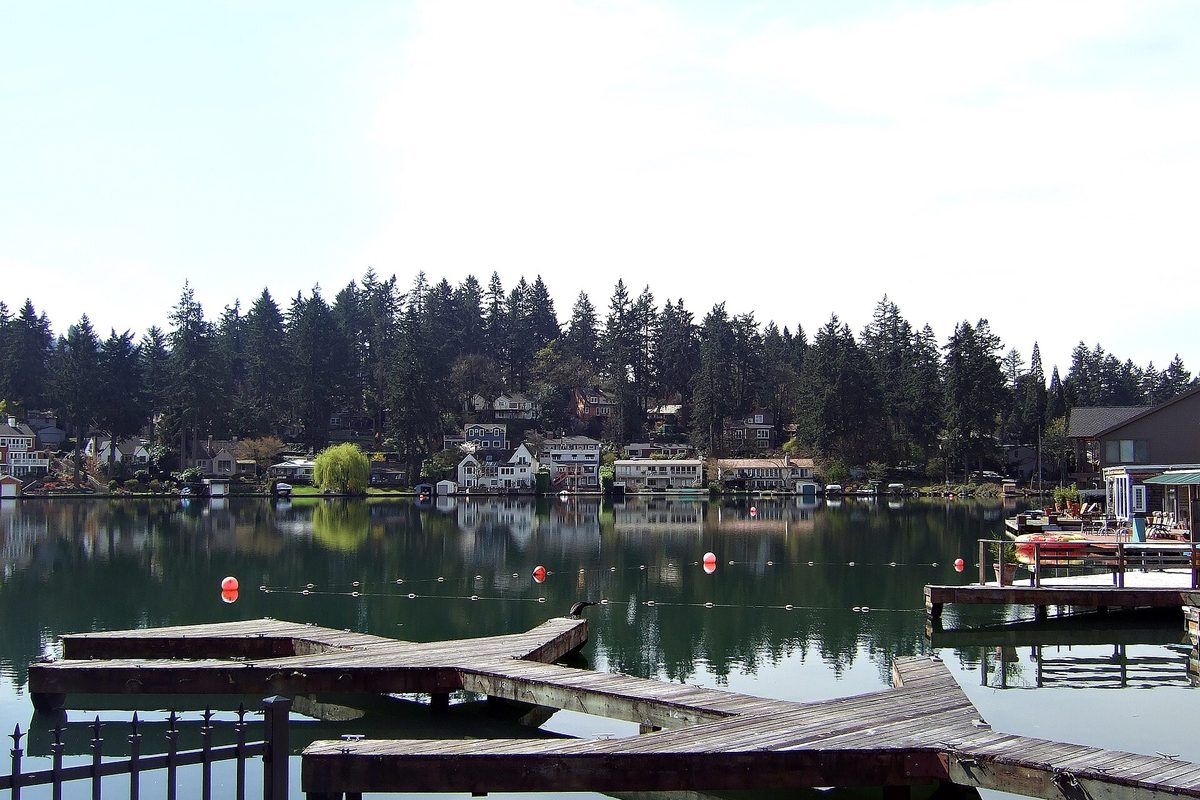 Weathered wooden docks on Lakewood Bay in Lake Oswego, with houses and tall evergreens reflected in the calm water