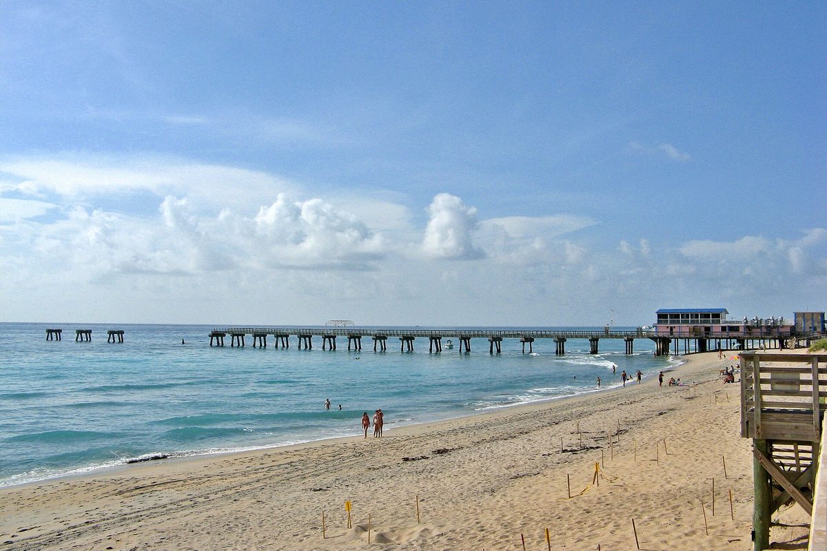 The Lake Worth Beach pier running out into blue-green Atlantic water, with a few swimmers on the sand and a building near the pier's end