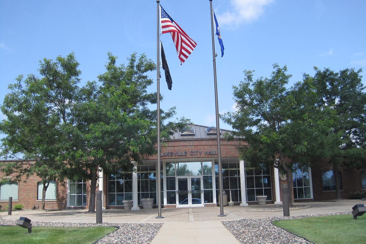 Lakeville City Hall, a brick building with tall glass entry windows, flags flying out front and green trees on both sides under a blue sky